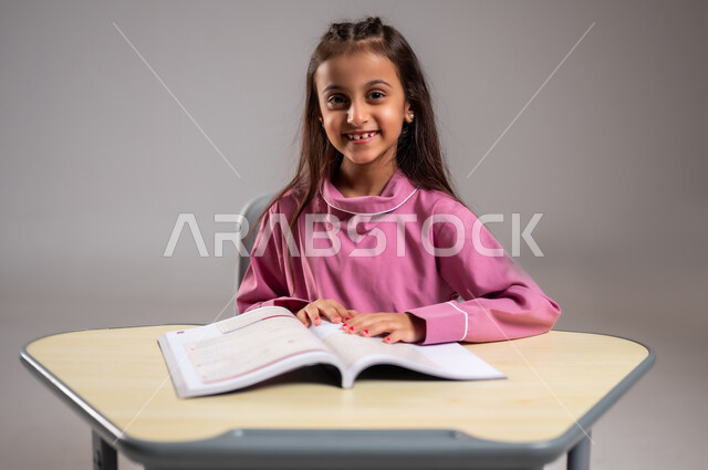 العودة إلى المدارس والطريق الى النجاح، أهمية العلم في تنمية القدرات العقلية، بورتريه مقرب لطالبة عربية سعودية خليجية تجلس على الكرسي مستعدة للدراسة، النظر الى الكاميرا بايماءات الفرح و الحماس بقدوم الفصل الدراسي، خلفية رمادية