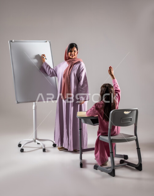مشاركة والتفاعل داخل الفصل الدراسي، التعابير وايماءات الحماس للاجابة على السؤال, بورتريه من الخلف لطالبة عربية خليجية سعودية ترفع يدها لتشارك في حل الاسئلة، التعليم في المملكة العربية السعودية، , خلفية رمادية