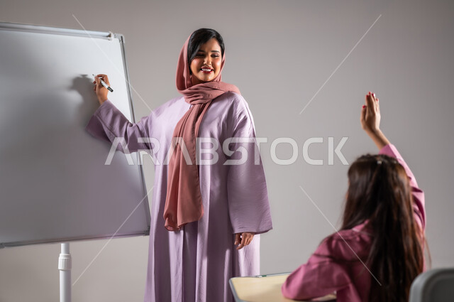 Achievement and success in the educational stages, a portrait of a veiled Saudi Arabian Gulf teacher wearing a colorful abaya explaining the lesson on the white school board, education according to the curricula prescribed in the Kingdom of Saudi Arabia, gray background