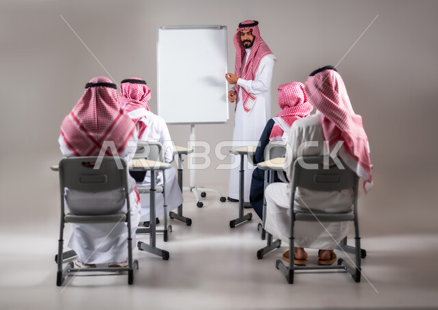 أهمية العلم في تنمية القدرات العقلية، المقررات و المناهج التعليمية في المملكة السعودية، بورتريه من الخلف لطلاب عرب خليجيين سعوديين يجلسون على المقعد المدرسي يستمعون الى استاذهم اثناء الدرس، ايماءات التركيز و التمعن بالدرس، استخدام اللوحة المدرسية البيضاء, خلفية رمادية