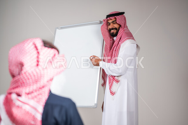 التربية و التعليم في المملكة العربية السعودية, ايماءات التركيز و التمعن, بورتريه مقرب لمعلم عربي خليجي سعودي يرتدي الثوب التقليدي يشرح لطالبه الدرس على اللوح، التعاون على فهم المسائل الرياضية، خلفية رمادية