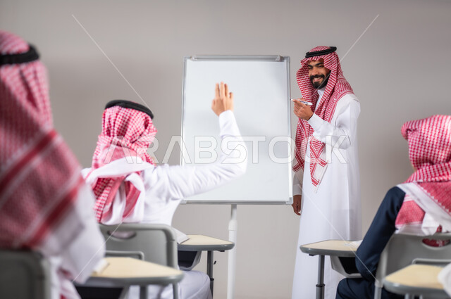 مشاركة الطلاب بواسطة رفع الايدي, التفاعل داخل الصف, استخدام اللوحة المدرسية البيضاء،  بورتريه لمعلم عربي خليجي سعودي يرتدي الثوب التقليدي يقوم بشرح الدرس، التربية والتعليم بالمنهاج المقررة، خلفية رمادية