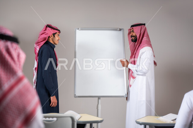 ايماءات التركيز و التمعن, التربية و التعليم في المملكة العربية السعودية، بورتريه لمعلم عربي خليجي سعودي يرتدي الثوب التقليدي يشرح لطالبه الدرس على اللوح في الصف المدرسي بحضور الطلاب، المشاركة الفعالة في الصف، خلفية رمادية