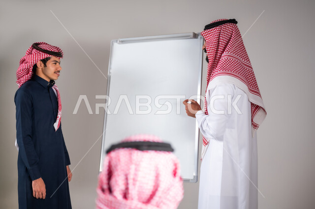 التربية و التعليم في المناهج المقررة، ايماءات التركيز و التمعن، بورتريه لمعلم عربي خليجي سعودي يرتدي الثوب التقليدي يشرح لطالبه مسألة رياضية على اللوح في الصف المدرسي، المشاركة الفعالة في الصف، خلفية رمادية