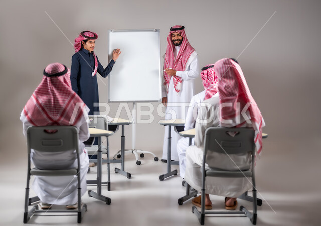الانجاز والنجاح في المرحلة التعليمية، ايماءات البشاشة والسرور، بورتريه لطالب عربي خليجي سعودي يرتدي الثوب التقليدي يشرح لزملائه الدرس على اللوح بمتابعة استاذه، المشاركة الفعالة في الصف، خلفية رمادية