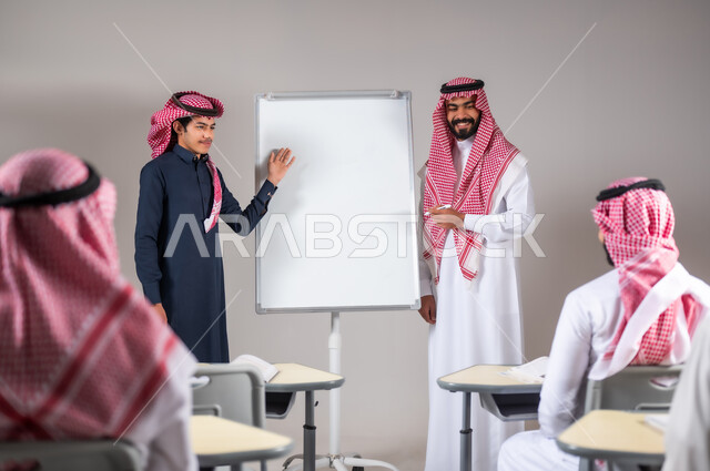 المشاركة الفعالة في الصف، بورتريه لطالب عربي خليجي سعودي يرتدي الثوب التقليدي و الشماغ يقوم بشرح الدرس لزملائه بإشراف معلمه، تلميذ متميز و متفوق في الدراسة, خلفية رمادية