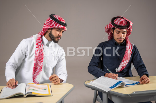 Active participation of students, performing homework in class, close-up portrait of two Saudi Gulf Arab students with gestures of contemplation and concentration in solving lesson problems, a Saudi Gulf Arab student cheating on his colleague in the classroom, education in the Kingdom of Saudi Arabia, gray background
