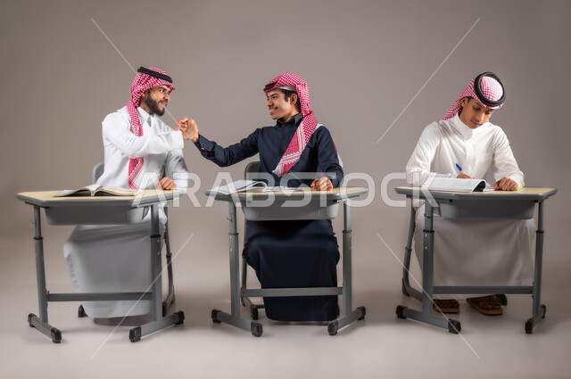 تماسك الايدي بتعابير النجاح و الاتفاق على شيئ ما, بورتريه لطالبان عربيان سعوديان خليجيان يرتديان الثوب التقليدي و الشماغ ينظران لبعضهما بايماءات الفرح و السرور, التدريس في المملكة العربية السعودية, خلفية رمادية