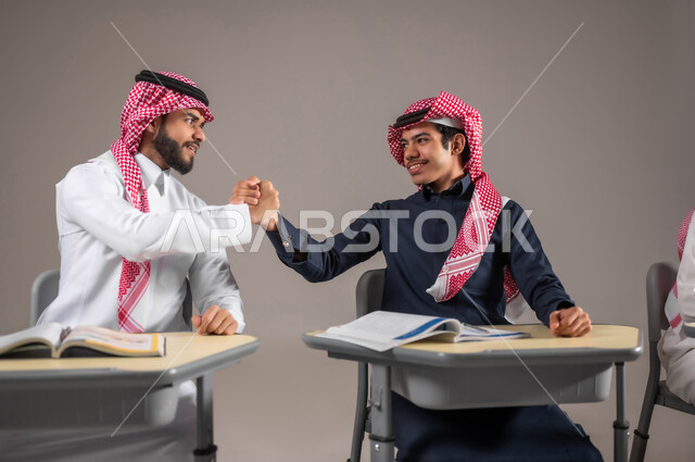 المشاركة و التعاون في الدراسة بين الطلاب, بورتريه لطالبان عربيان سعوديان خليجيان يرتديان الثوب التقليدي و الشماغ يمسكان قضبة يديهما بايماءات الصداقة و الاخوة, تماسك الايدي بتعابير النجاح و الاتفاق على شيئ ما, خلفية رمادية