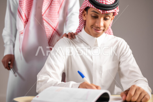 الدراسة و الاجتهاد من اجل التفوق، التربية و التعليم في المملكة العربية السعودية، بورتريه مقرب لمعلم عربي خليجي سعودي يرتدي الثوب التقليدي يقوم بمراقبة الطالب عن قرب اثناء تقديم اختبارات نهاية الفصل الدراسي، يضع يده على كتف الطالب، خلفية رمادية