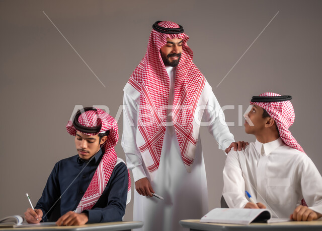 الانجاز والنجاح في المرحلة التعليمية، أهداف التربية والتعليم بالمناهج المقررة للطلاب، امتحانات نهاية الفصل الدراسي، بورتريه مقرب لمعلم عربي خليجي سعودي يرتدي الثوب التقليدي يضع يده على كتف الطالب، مراقبة التلاميذ عن قرب اثناء تقديم الاختبارات، خلفية رمادية