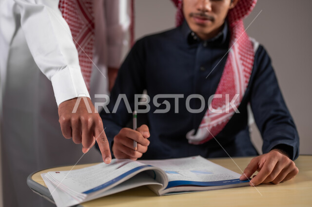 مراقبة التلاميذ عن قرب اثناء تقديم الاختبارات، امتحانات نهاية الفصل الدراسي، النجاح في المرحلة التعليمية، أهداف التربية و التعليم بالمناهج المقررة للطلاب، بورتريه مقرب لمعلم عربي خليجي سعودي يرتدي الثوب التقليدي يشير باصبع السبابة الى شيء ما، خلفية رمادية