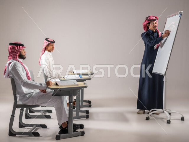 الانجاز و النجاح في المرحلة التعليمية، بورتريه لطالب عربي خليجي سعودي يرتدي الثوب التقليدي يقوم بشرح الدرس لزملاءه عن طريق استخدام اللوحة المدرسية البيضاء، المشاركة الفعالة في الصف، التربية والتعليم بالمنهاج المقررة، خلفية رمادية