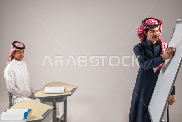 المشاركة الفعالة في الصف، الانجاز و النجاح في المرحلة التعليمية، بورتريه لطالب عربي خليجي سعودي يرتدي الثوب التقليدي يقوم بشرح الدرس لزملاءه عن طريق استخدام اللوحة المدرسية البيضاء، التربية والتعليم بالمنهاج المقررة، خلفية رمادية