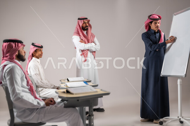 الانجاز و التفوق في المرحلة التعليمية، التربية و التعليم بالمنهاج المقررة، المشاركة الفعالة في الصف، بورتريه لطالب عربي خليجي سعودي يرتدي الثوب التقليدي يقوم بالشرح والإجابة على سؤال المعلم امام التلاميذ، استخدام اللوحة المدرسية البيضاء، خلفية رمادية