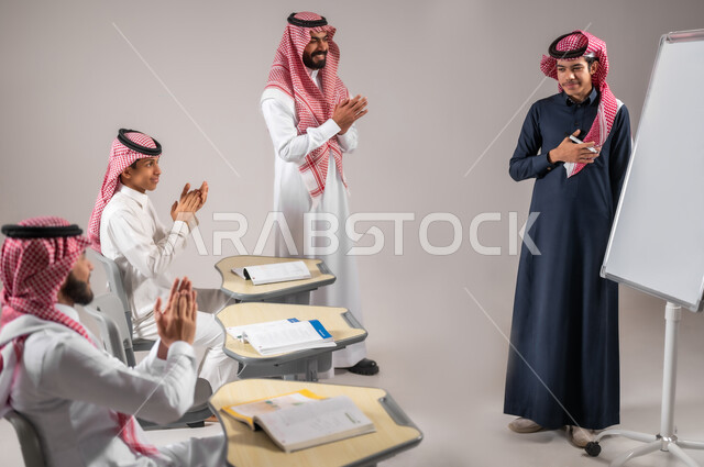 ايماءات التشجيع  و التصفيق داخل الفصل الدراسي، بورتريه لطالب عربي خليجي سعودي يرتدي الثوب التقليدي يقوم بالشرح والإجابة على سؤال المعلم امام التلاميذ، استخدام اللوحة المدرسية البيضاء، المشاركة الفعالة في الصف، التربية والتعليم بالمنهاج المقررة، خلفية رمادية