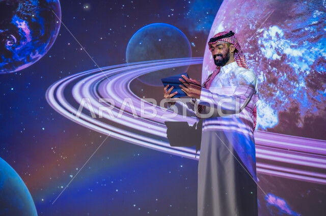 ايماءات السعادة والفخر، رؤية مستقبلية سعودية ٢٠٣٠، نمو قطاع الفضاء في المملكة العربية السعودية، التطور الرقمي والتكنولوجي، شاب عربي سعودي خليجي مبتسم يرتدي الثوب التقليدي يقف بين الكواكب والنجوم يحمل بيده جهاز لوحي، خلفية هيولوجرامية ثلاثية الأبعاد للكواكب