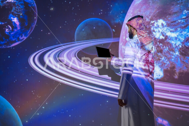 رؤية مستقبلية سعودية ٢٠٣٠، نمو قطاع الفضاء في المملكة العربية السعودية، التطور الرقمي والتكنولوجي، شاب عربي سعودي خليجي يرتدي الثوب التقليدي يقف بين الكواكب والنجوم يحمل بيده جهاز لوحي، عرض شاشة سوداء فارغة، خلفية هيولوجرامية ثلاثية الأبعاد  للكواكب