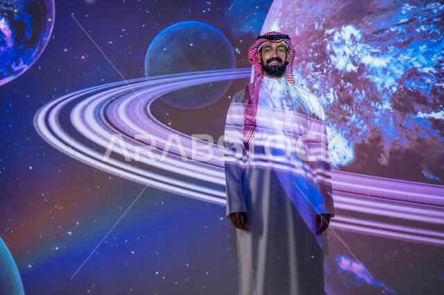 نمو قطاع الفضاء في المملكة العربية السعودية، ايماءات السعادة والسرور، التطور الرقمي والتكنولوجي، شاب عربي سعودي خليجي مبتسم يرتدي الثوب التقليدي يقف بين الكواكب والنجوم ينظر إلى الكاميرا، رؤية مستقبلية سعودية ٢٠٣٠، خلفية هيولوجرامية ثلاثية الأبعاد للكواكب