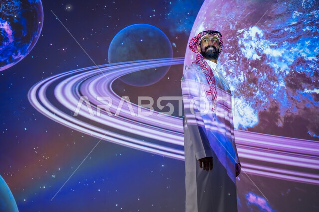 ايماءات السعادة والسرور، التطور الرقمي والتكنولوجي، نمو قطاع الفضاء في المملكة العربية السعودية، شاب عربي سعودي خليجي مبتسم يرتدي الثوب التقليدي يقف بين الكواكب والنجوم ينظر إلى الأعلى، رؤية مستقبلية سعودية ٢٠٣٠، خلفية هيولوجرامية ثلاثية الأبعاد للكواكب