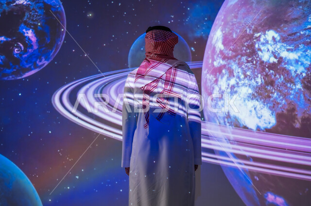 التطور الرقمي والتكنولوجي، نمو قطاع الفضاء في المملكة العربية السعودية، شاب عربي سعودي خليجي يرتدي الثوب التقليدي ينظر إلى الكواكب والنجوم، رؤية مستقبلية سعودية ٢٠٣٠، خلفية هيولوجرامية ثلاثية الأبعاد للكواكب