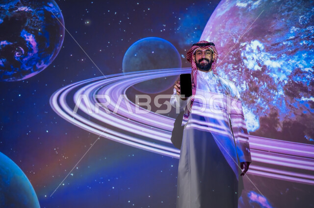 رحلة السعودية نحو الفضاء الخارجي، رؤية مستقبلية 2030، رجل سعودي عربي خليجي مبتسم يقف بين الكواكب والنجوم يحمل بيده الهاتف المحمول، ايماءات السعادة والفخر، النمو والتطوير لقطاع الفضاء في المملكة العربية السعودية، عرض شاشة الجوال السوداء الفارغة