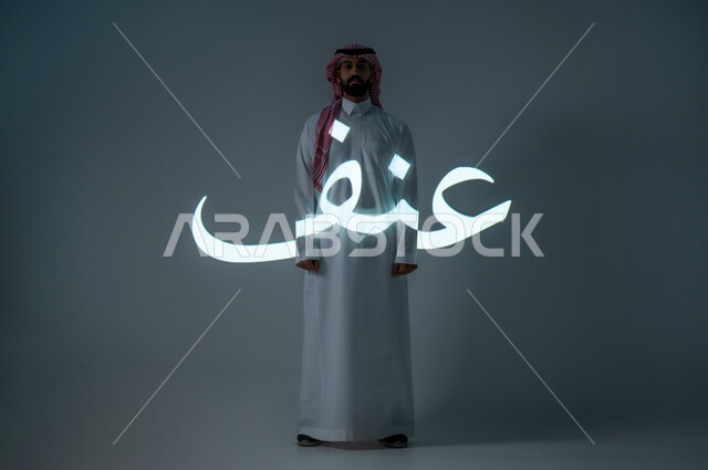 مخطوطة كلمة عنف باللغة العربية، بورتريه شاب عربي خليجي سعودي يرتدي الثوب التقليدي ويضع الشماغ، الطول الكامل للجسم، خلفية رمادية