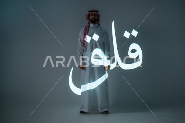 مخطوطة كلمة قلق باللغة العربية، بورتريه شاب عربي خليجي سعودي يرتدي الثوب التقليدي ويضع الشماغ، الطول الكامل للجسم، خلفية رمادية