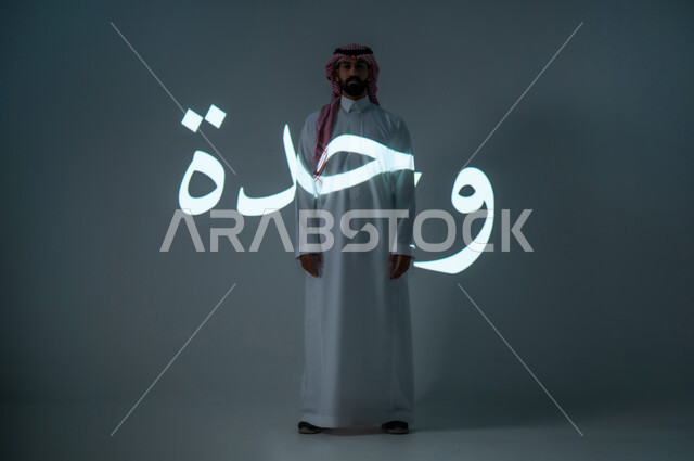 مخطوطة كلمة وحدة باللغة العربية، بورتريه شاب عربي خليجي سعودي يرتدي الثوب التقليدي ويضع الشماغ، الطول الكامل للجسم، خلفية رمادية