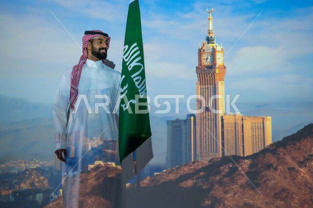 التعبير عن الحب والولاء في يوم العلم 11 مارس, الاحتفال بذكرى اليوم الوطني 23 سبتمبر, شاب عربي خليجي سعودي مبتسم يرتدي الزي التقليدي يقف بجانب علم المملكة العربية السعودية، الخلفية برج الساعة الملكي في الحرم المكي الشريف