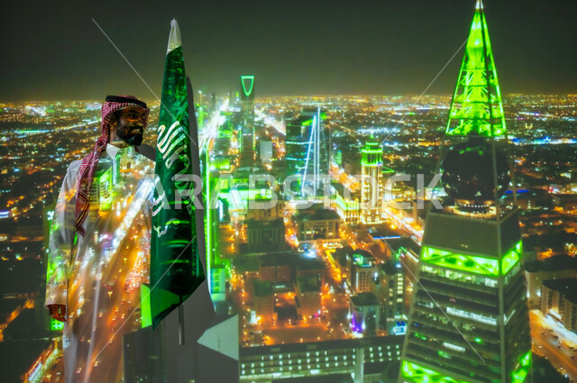 ذكرى اليوم الوطني للسعودية في 23 سبتمبر، الاحتفال بالأعياد والمناسبات الوطنية، رجل عربي خليجي سعودي مبتسم يرتدي الزي التقليدي يقف بجانب علم المملكة العربية السعودية يحتفل يوم العلم السعودي في11 مارس، الخلفية برج الفيصلية في الرياض