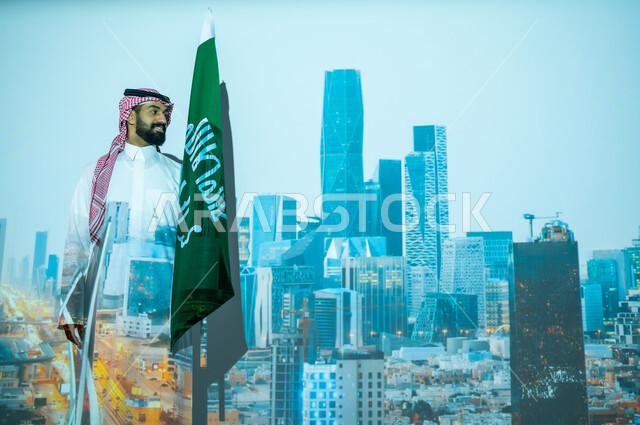 الاحتفال بيوم العلم السعودي في11 مارس، ذكرى اليوم الوطني للسعودية في 23 سبتمبر، الأعياد والمناسبات الوطنية، رجل عربي خليجي سعودي مبتسم يرتدي الزي التقليدي ينظر إلى الكاميرا ويقف بجانب علم المملكة العربية السعودية، الخلفية مركز الملك عبدالله المالي كافد في الرياض