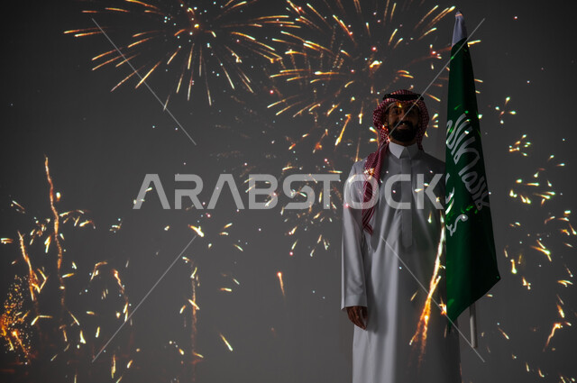 Celebrating National Day, September 23, participating in recreational activities and events, a smiling Saudi Gulf Arab young man wearing traditional clothing standing next to the Kingdom’s flag and looking at the camera, commemorating Saudi Flag Day on March 11, enjoying firecrackers and fireworks on national holidays and occasions