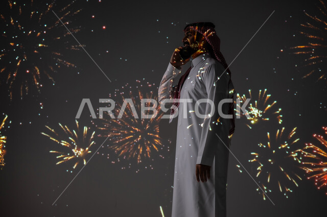 الاحتفالات والفعاليات الترفيهية، الاستمتاع بالمفرقعات والألعاب النارية في الأعياد والمناسبات، شاب عربي خليجي سعودي مبتسم يرتدي الزي التقليدي يستخدم الهاتف المحمول لتبادل التهاني مع العائلة والأصدقاء، التواصل عن بعد من خلال الأجهزة الالكترونية
