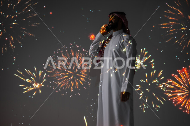الاستمتاع بالمفرقعات والألعاب النارية في الأعياد والمناسبات، المشاركة بالأنشطة الترفيهية والفعاليات الاحتفالية، شاب عربي خليجي سعودي مبتسم يرتدي الزي التقليدي يستخدم الهاتف المحمول لتبادل التهاني مع العائلة والأصدقاء، التواصل عن بعد من خلال الأجهزة الالكترونية