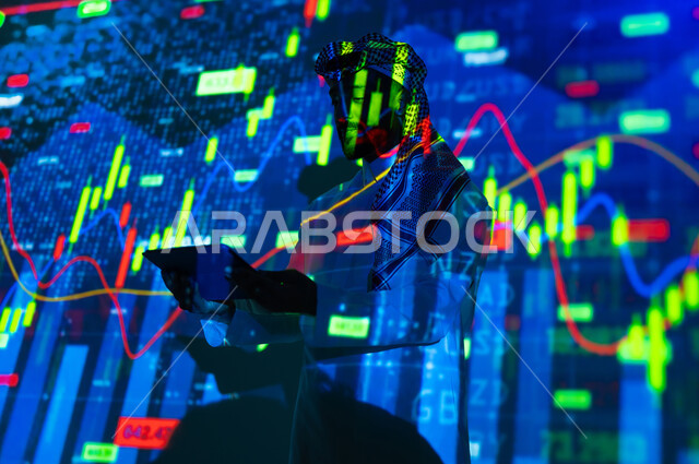 إحصائيات وبيانات البورصة وسوق الأسهم, استخدام التقنيات لمراقبة الاقتصاد و معلومات الاستثمارات المالية, رجل عربي خليجي سعودي يرتدي الثوب التقليدي و الشماغ يشير بإصبع السبابة الى شاشة جهاز التابلت يقف أمام شاشة مليئة بالأرقام, خلفية هولوجرامية ثلاثية الأبعاد