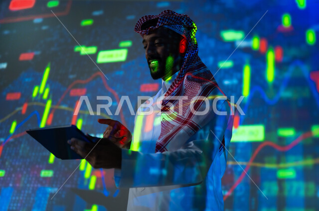 إحصائيات وبيانات البورصة وسوق الأسهم, استخدام التقنيات لمراقبة الاقتصاد و معلومات الاستثمارات المالية, رجل عربي خليجي سعودي يرتدي الثوب التقليدي و الشماغ يشير بإصبع السبابة الى شاشة جهاز التابلت يقف أمام شاشة مليئة بالأرقام, خلفية هولوجرامية ثلاثية الأبعاد
