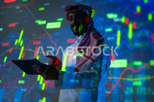 استخدام التقنيات لمراقبة الاقتصاد و معلومات الاستثمارات المالية, جمع بيانات البورصة وسوق الأسهم, رجل عربي خليجي سعودي يرتدي الثوب التقليدي و الشماغ ينظر الى الكاميرا يمسك بيده جهاز التابلت يقف أمام شاشة مليئة بالأرقام, خلفية هولوجرامية ثلاثية الأبعاد