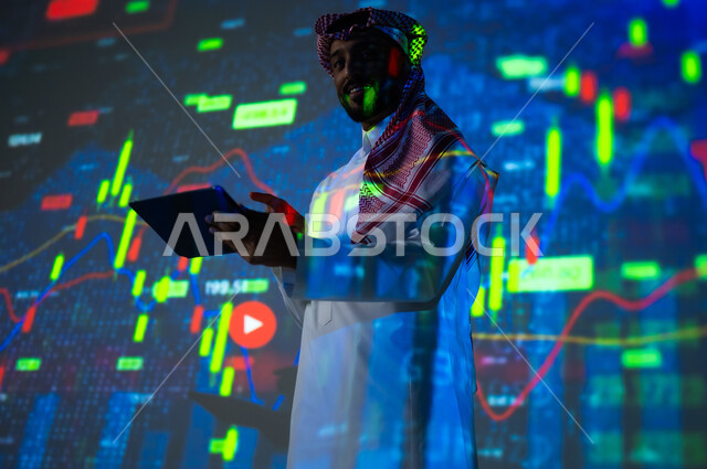 استخدام التقنيات لمراقبة الاقتصاد و معلومات الاستثمارات المالية, جمع بيانات البورصة وسوق الأسهم, رجل عربي خليجي سعودي يرتدي الثوب التقليدي و الشماغ ينظر الى الكاميرا يمسك بيده جهاز التابلت يقف أمام شاشة مليئة بالأرقام, خلفية هولوجرامية ثلاثية الأبعاد