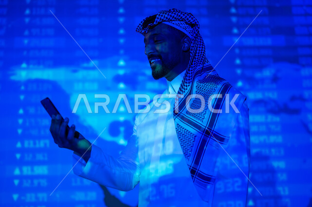 التغيرات العالمية بأسعار أسهم البورصة , صورة من الخلف لرجل عربي خليجي سعودي يرتدي الثوب التقليدي و الشماغ يقف أمام شاشة مليئة بالأرقام و الإحصائيات يمسك بيده الهاتف المحمول, خلفية هولوجرامية ثلاثية الأبعاد لخريطة العالم باللون الأزرق