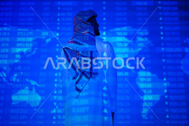 التغيرات العالمية بأسعار أسهم البورصة , صورة من الخلف لرجل عربي خليجي سعودي يرتدي الثوب التقليدي و الشماغ يقف أمام شاشة مليئة بالأرقام و الإحصائيات, خلفية هولوجرامية ثلاثية الأبعاد لخريطة العالم باللون الأزرق