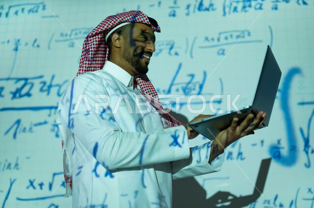 استخدام التقنيات الحديثة, دمج العلم بالتكنولوجيا، أرقام وحسابات في مادة الرياضيات، رجل عربي خليجي سعودي يرتدي الثوب التقليدي والشماغ يحمل بيده جهار اللابتوب المحمول , التطور العلمي و التقني في المملكة العربية السعودية, خلفية قوانين ومعادلات رياضية