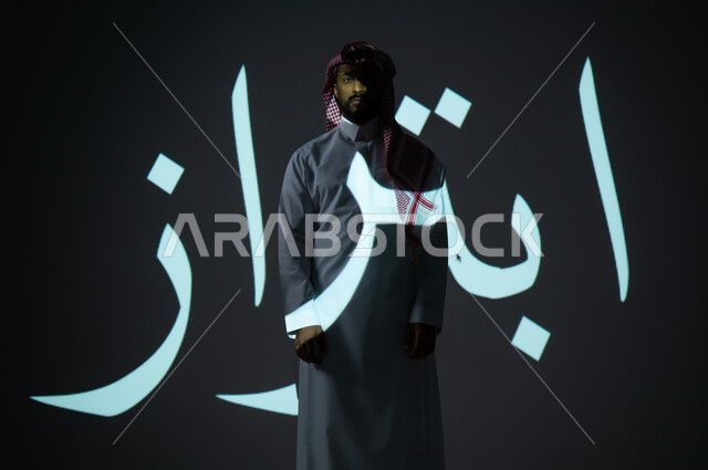 مخطوطة كلمة ابتزاز باللغة العربية، بورتريه شاب عربي خليجي سعودي يرتدي الثوب التقليدي ويضع الشماغ يقف أمام الكلمة، الطول الكامل للجسم، خلفية رمادية