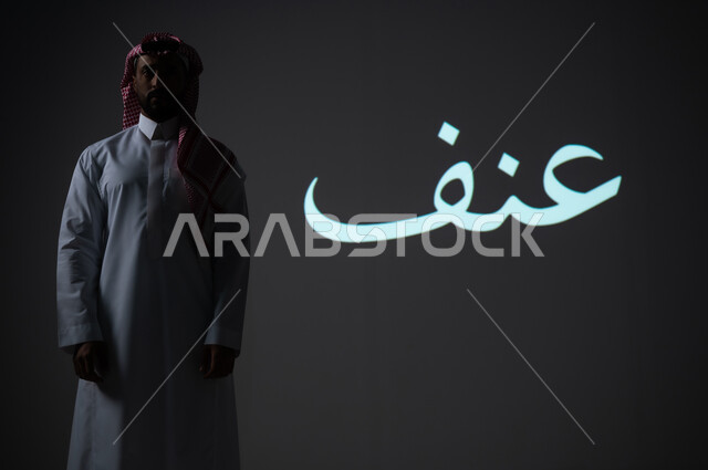 مخطوطة كلمة عنف باللغة العربية، بورتريه شاب عربي خليجي سعودي يرتدي الثوب التقليدي ويضع الشماغ يقف بجانب الكلمة، الطول الكامل للجسم، خلفية رمادية