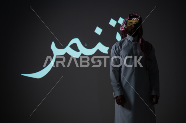 مخطوطة كلمة تنمر باللغة العربية، بورتريه شاب عربي خليجي سعودي يرتدي الثوب التقليدي ويضع الشماغ يقف بجانب الكلمة ينظر إلى الكاميرا، الطول الكامل للجسم، خلفية رمادية