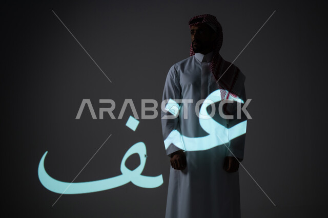 مخطوطة كلمة عنف باللغة العربية، بورتريه شاب عربي خليجي سعودي يرتدي الثوب التقليدي ويضع الشماغ يقف بجانب الكلمة، الطول الكامل للجسم، خلفية رمادية