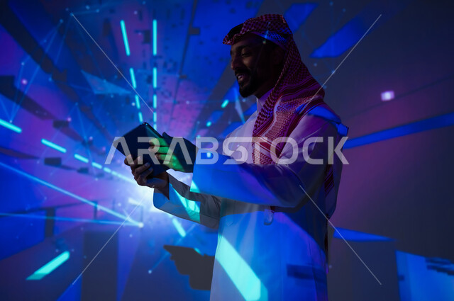 رؤية مستقبلية ٢٠٣٠، الاهتمام بالقطاع الالكتروني في المملكة العربية السعودية، تعزيز مفهوم التقدم التكنولوجي والتطور في المجتمع السعودي، التقنيات الحديثة والمتطورة، شاب عربي خليجي سعودي يرتدي الثوب التقليدي يمسك بيده جهاز لوحي، الخلفية إضاءة النيون الزرقاء