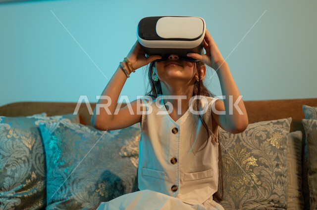 Expressions of the face and hands indicating enthusiasm, technical and technological progress and development, amusement, enjoyment, and interaction with electronic games, watching 3D cartoon films through modern virtual reality glasses, a Saudi Arabian Gulf girl wearing imaginary world glasses.