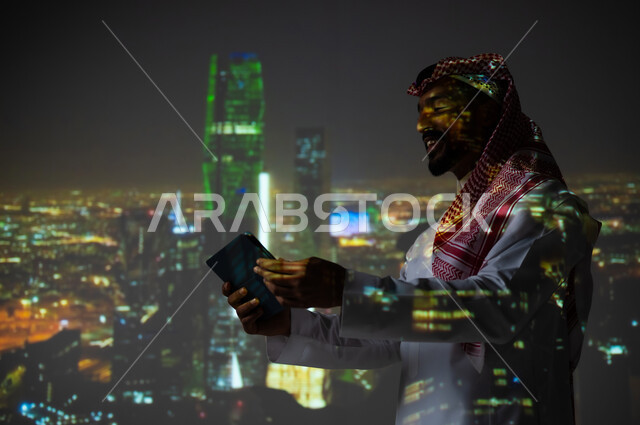 يوم العلم السعودي 11 مارس، الاحتفال باليوم الوطني 23 سبتمبر في المملكة العربية السعودية، رجل عربي خليجي سعودي يرتدي الثوب التقليدي يمسك بيده جهاز التابلت، الوقوف أمام مركز الملك عبدلله المالي في الرياض, الأبراج وناطحات السحاب المضيئة باللون الأخضر، رؤية مستقبلية سعودية 2030