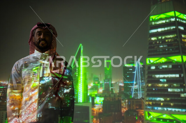ايماءات السعادة والفخر، معالم سياحية مشهورة، الفن الهندسي المعماري المتميز، شاب عربي خليجي سعودي يرتدي الثوب التقليدي يقف أمام الأبراج وناطحات السحاب المضيئة باللون الأخضر ليلا ينظرإلى الكاميرا، النمو والتقدم والتطور العمراني في المملكة العريبة السعودية